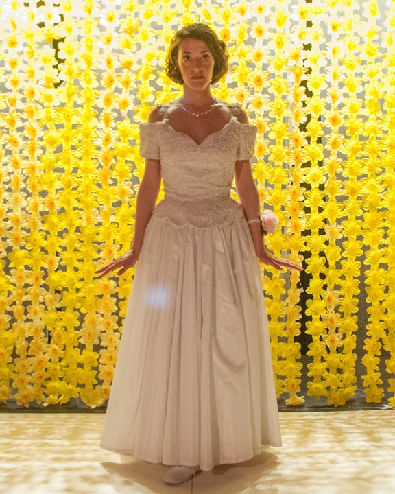 A debutante in a white gown stands before a wall of yellow flowers in Debutante.